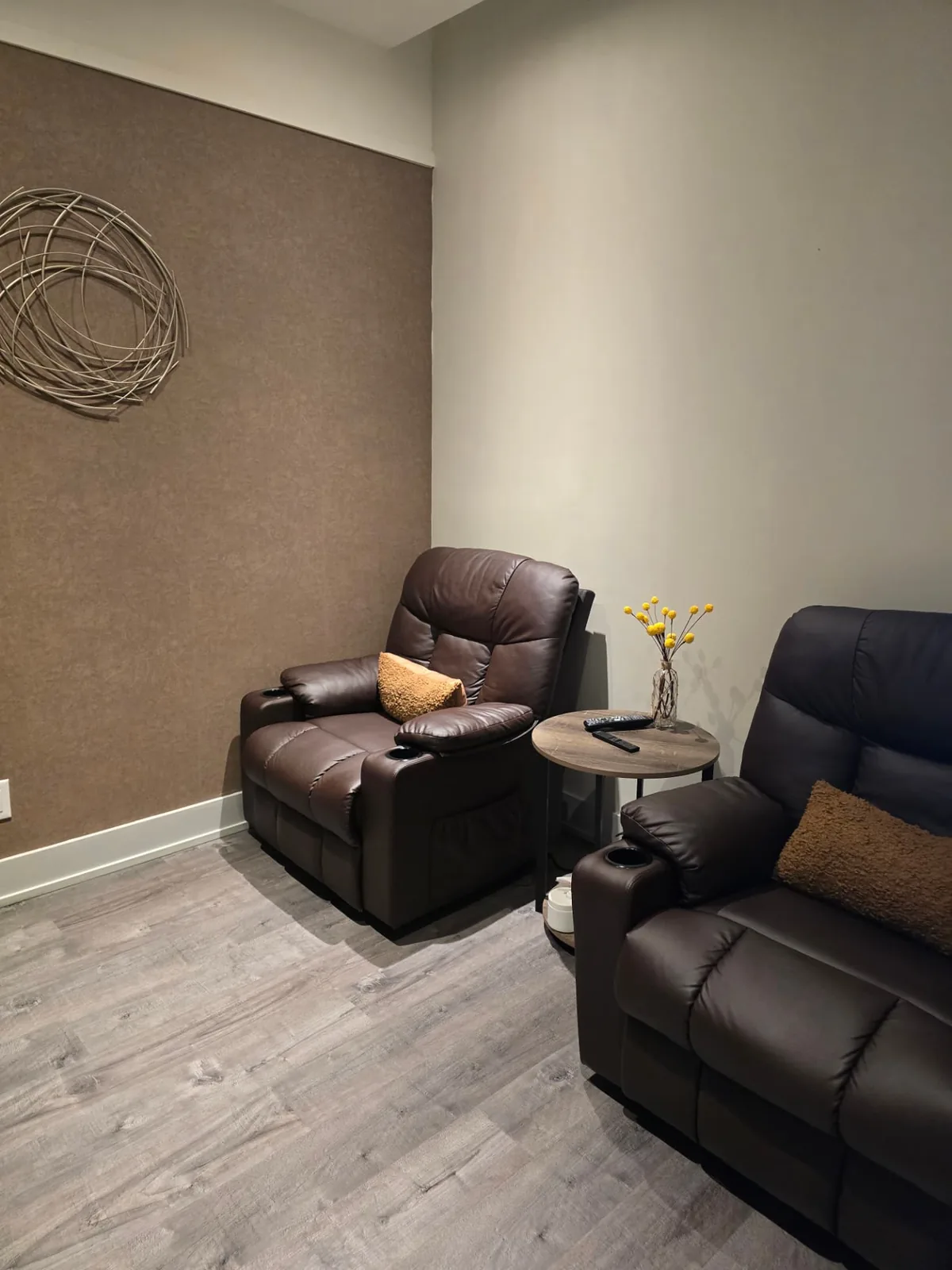 A quiet corner with two brown leather reclining chairs, a small round table and a vase of yellow blooms.