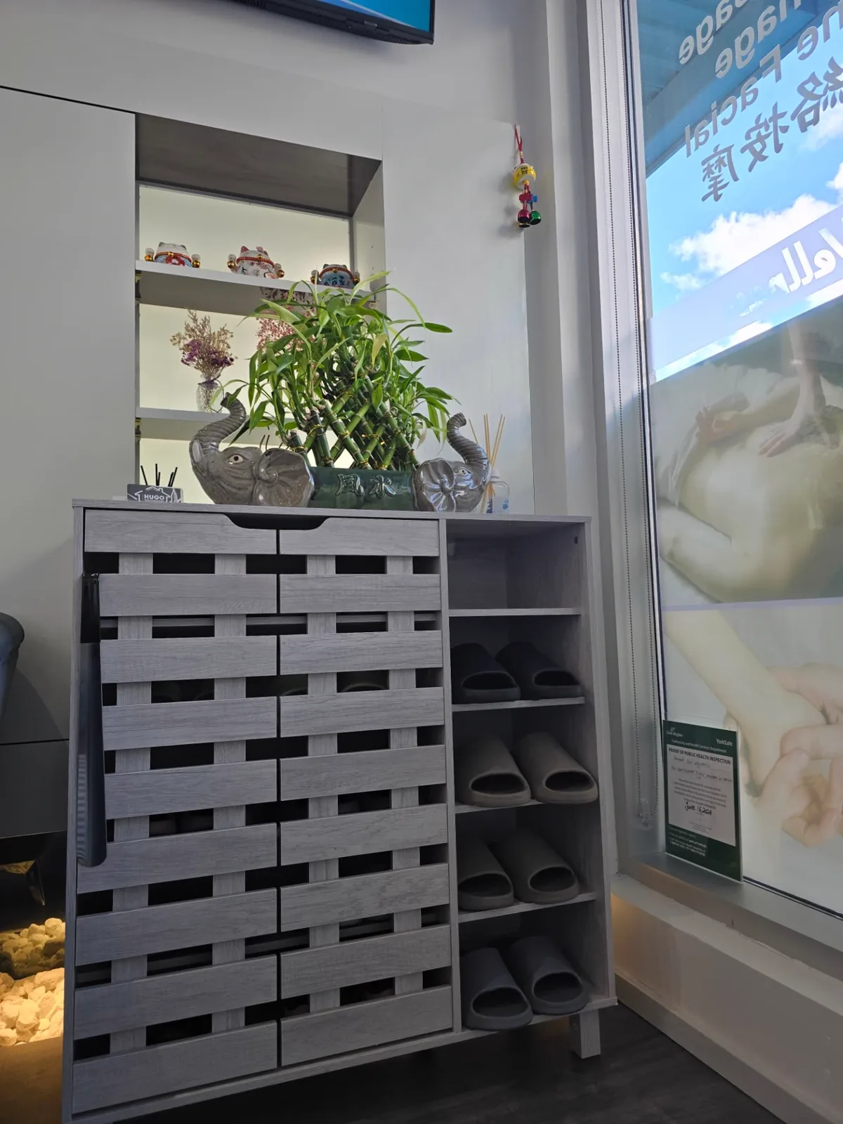 A wooden cabinet of fresh grey slippers by the window, topped with bamboo and ceramic elephants.