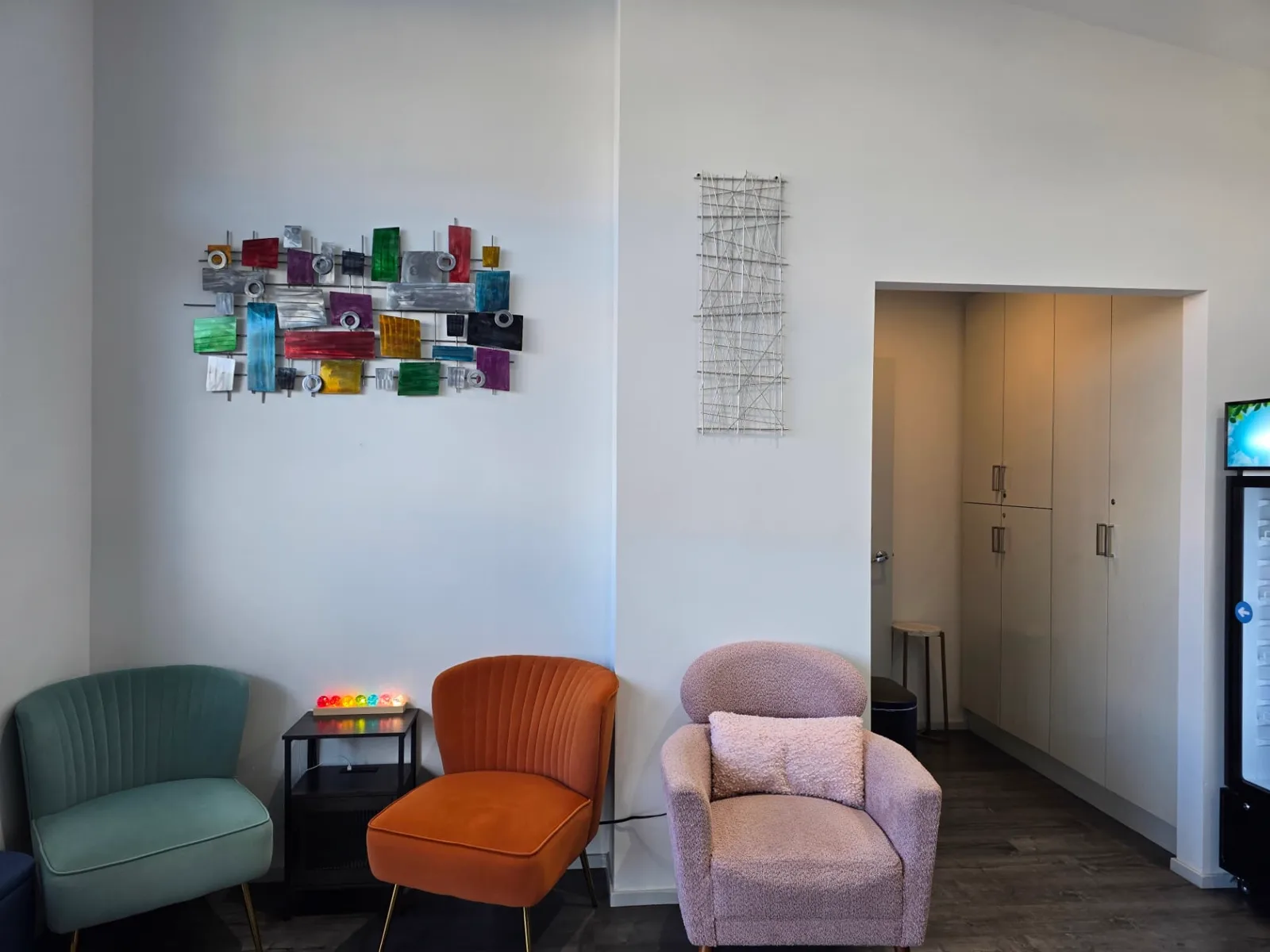 A waiting corner at Radiant Sun Wellness with green, orange and pink armchairs beneath a colourful glass wall sculpture.